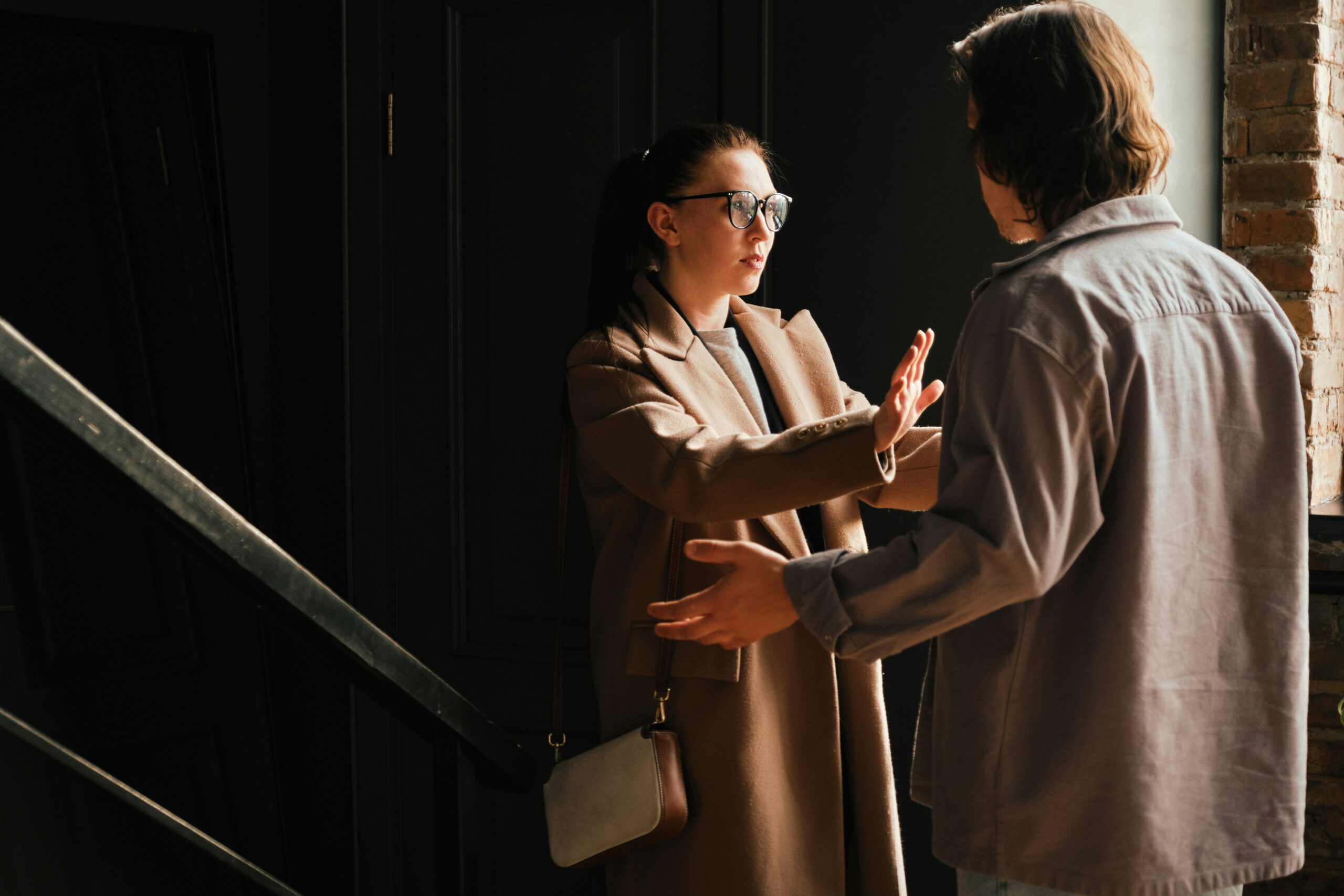 A woman wearing glasses and a brown coat raises her hands to stop a man in a gray shirt, appearing to refuse or argue with him, possibly signaling tension from an abusive relationship while standing indoors near stairs and a brick wall.