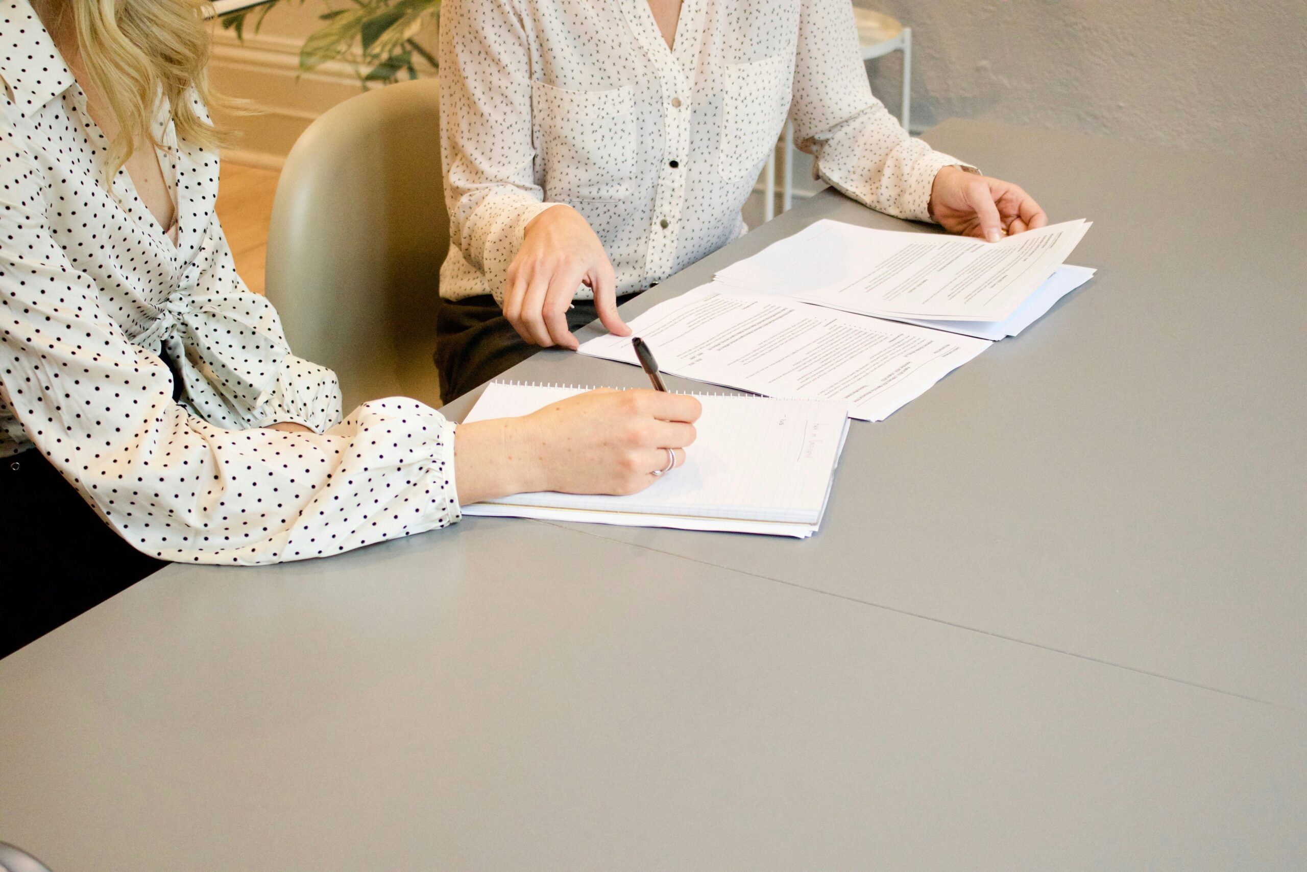 Two women in polka dot blouses sit at a table reviewing and writing on documents, with one woman pointing at a page and the other holding a pen. The focus is on estate planning, their hands and papers.