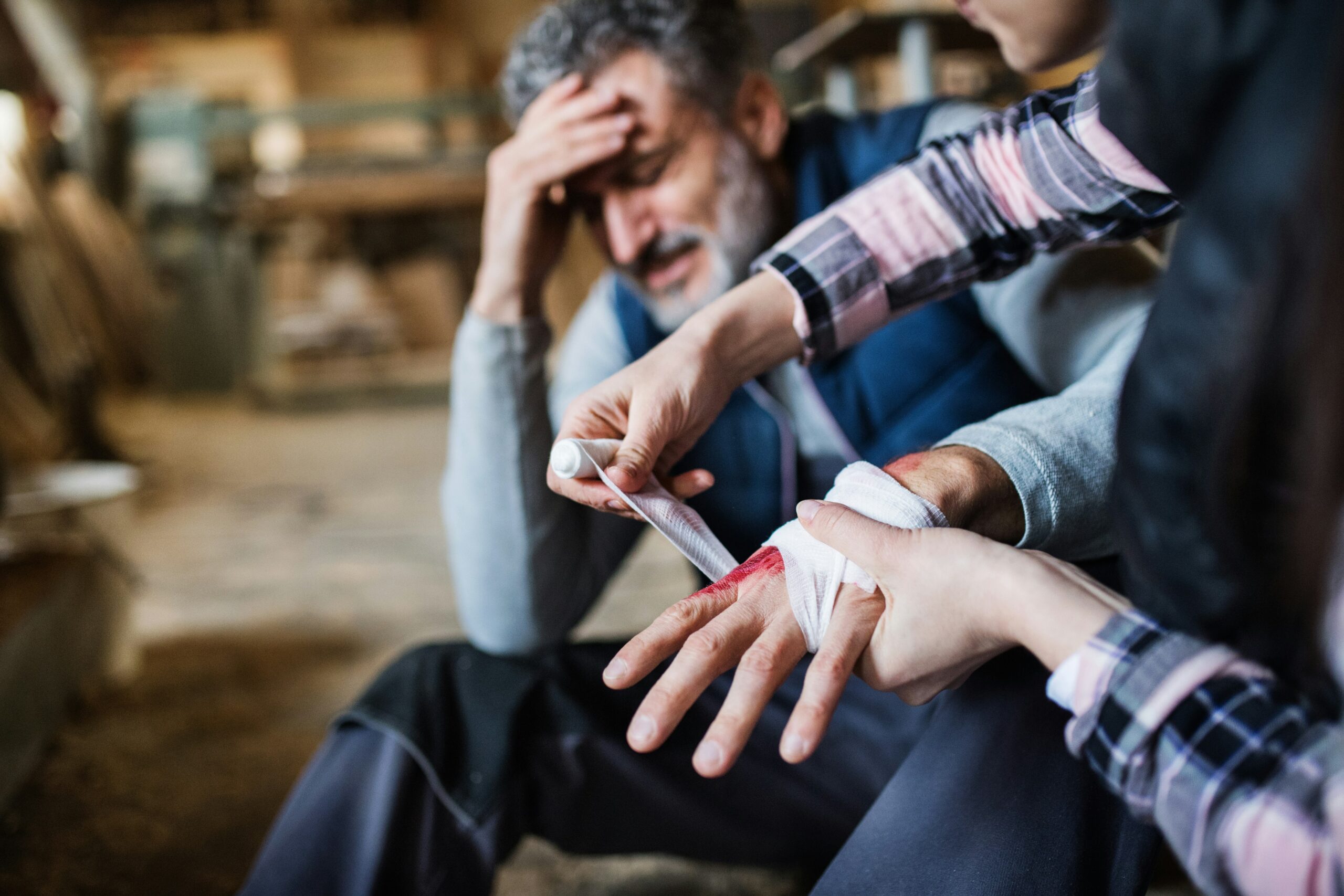 A person is wrapping a bandage around another personal injury case, bleeding hand. The injured person looks distressed, sitting with one hand on their forehead in a workshop or industrial setting.