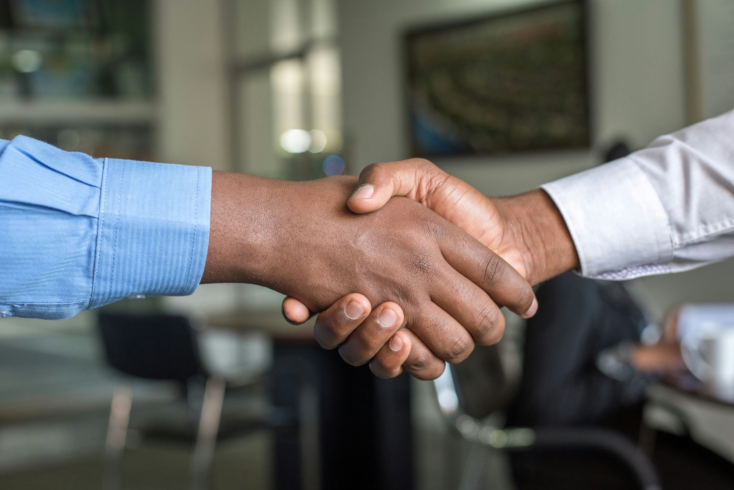 Two people wearing long-sleeved shirts are shaking hands in an office setting, symbolizing agreement or partnership. The background is blurred with office furniture visible.