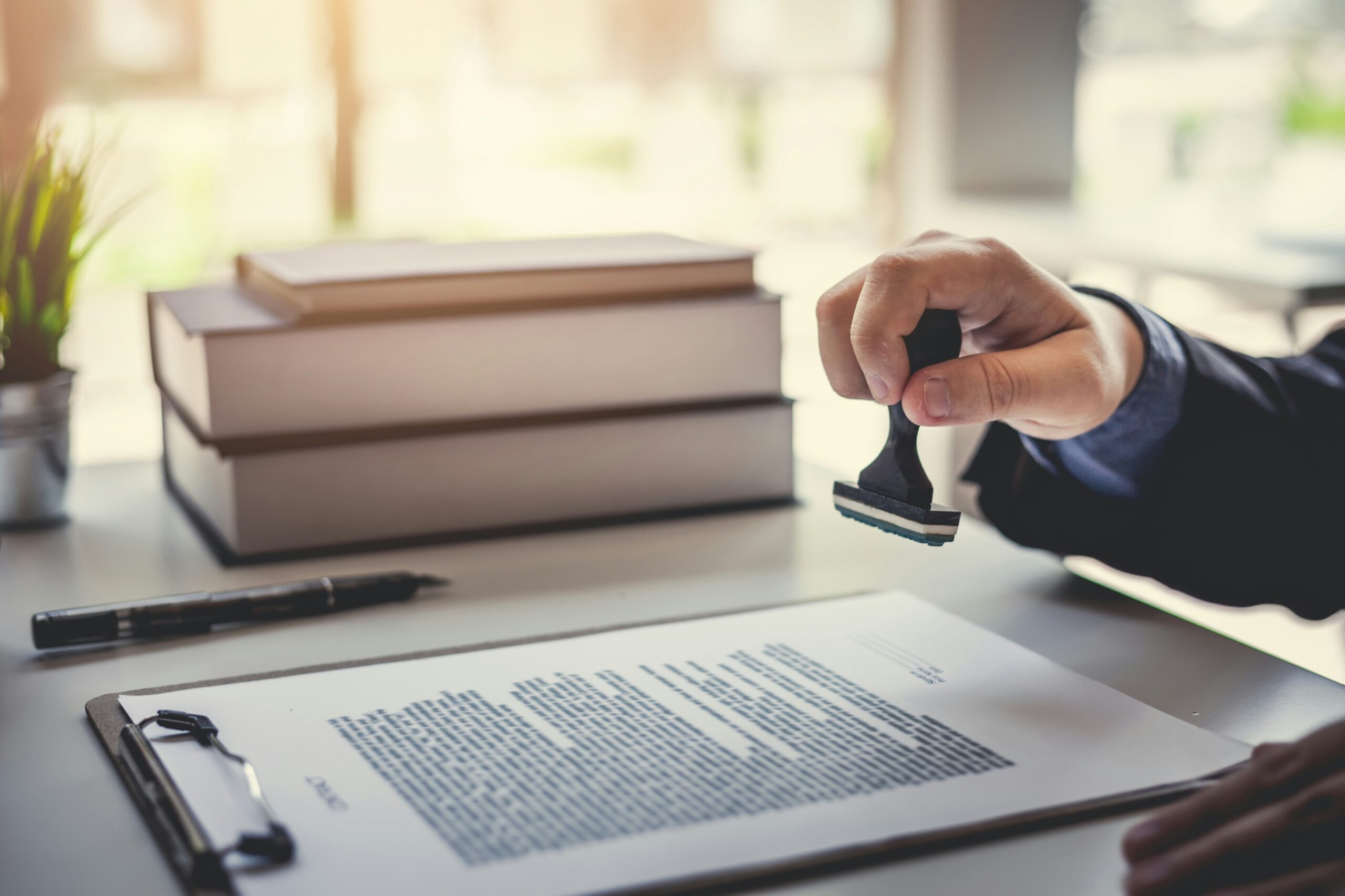 A person’s hand holding a rubber stamp above a document on a clipboard, with books, a pen, and a small potted plant on a desk in the background.