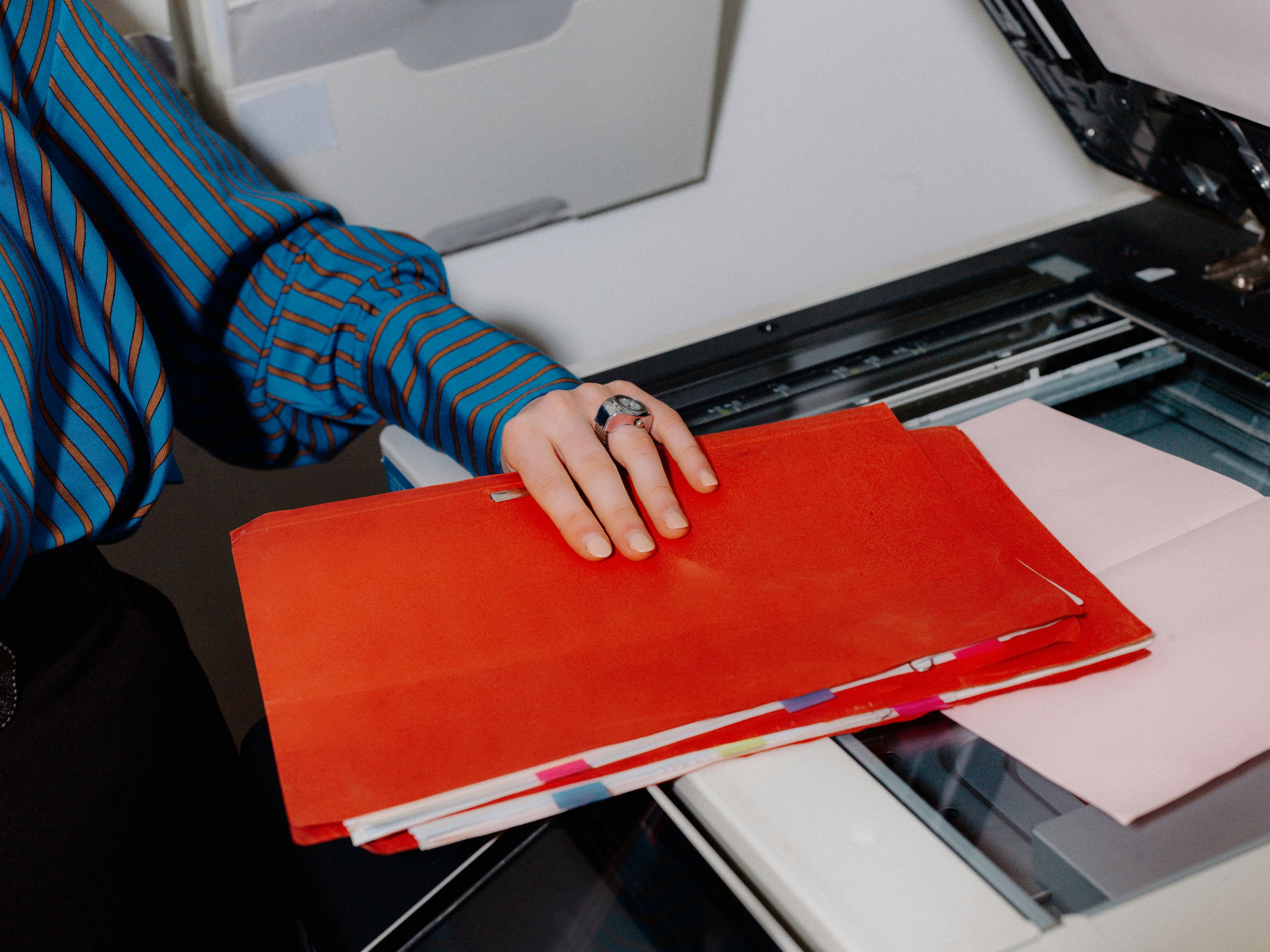 A person wearing a blue striped shirt holds a red folder while using a photocopier. Pink sheets of paper are on the copier glass. Only their hand and part of their body are visible.