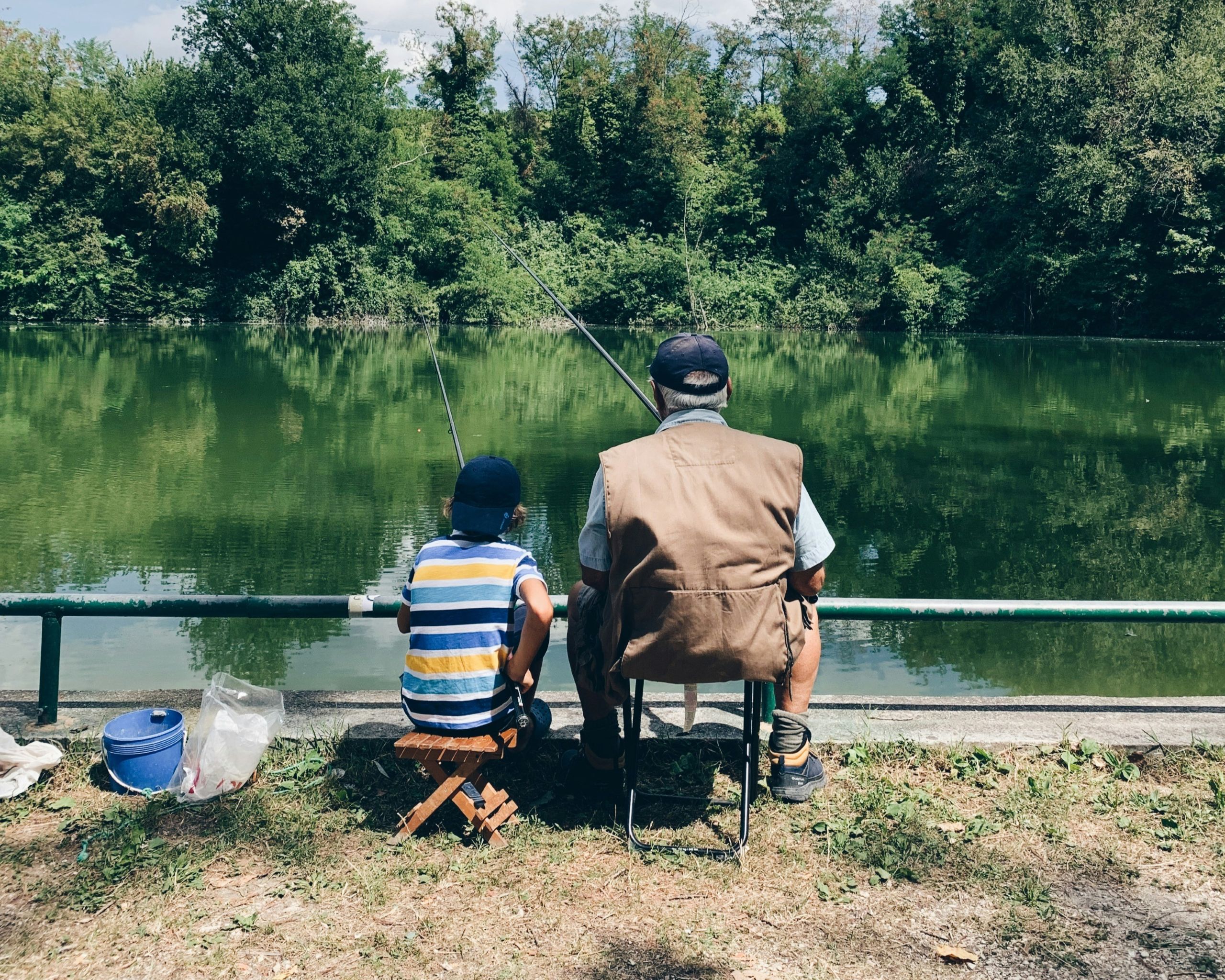 An older man and a young boy, perhaps enjoying time together amid child custody arrangements, sit side by side fishing at a calm, green lake. They both wear hats and face the water with fishing poles in hand, supplies resting on the ground nearby.