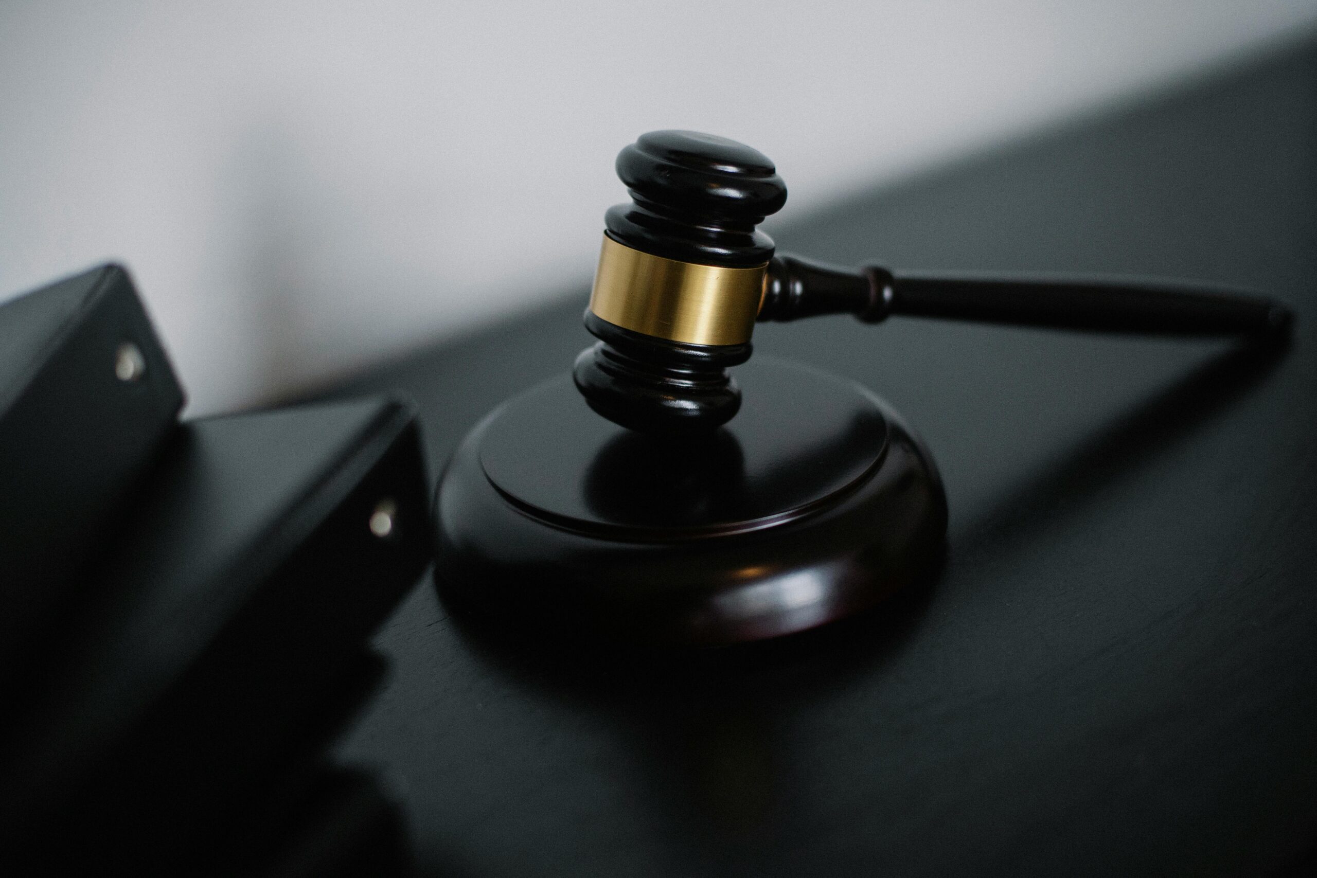 A wooden judge’s gavel with a gold band rests on a sound block atop a dark surface, suggesting themes of business compliance, with part of two black folders visible on the left side of the image.