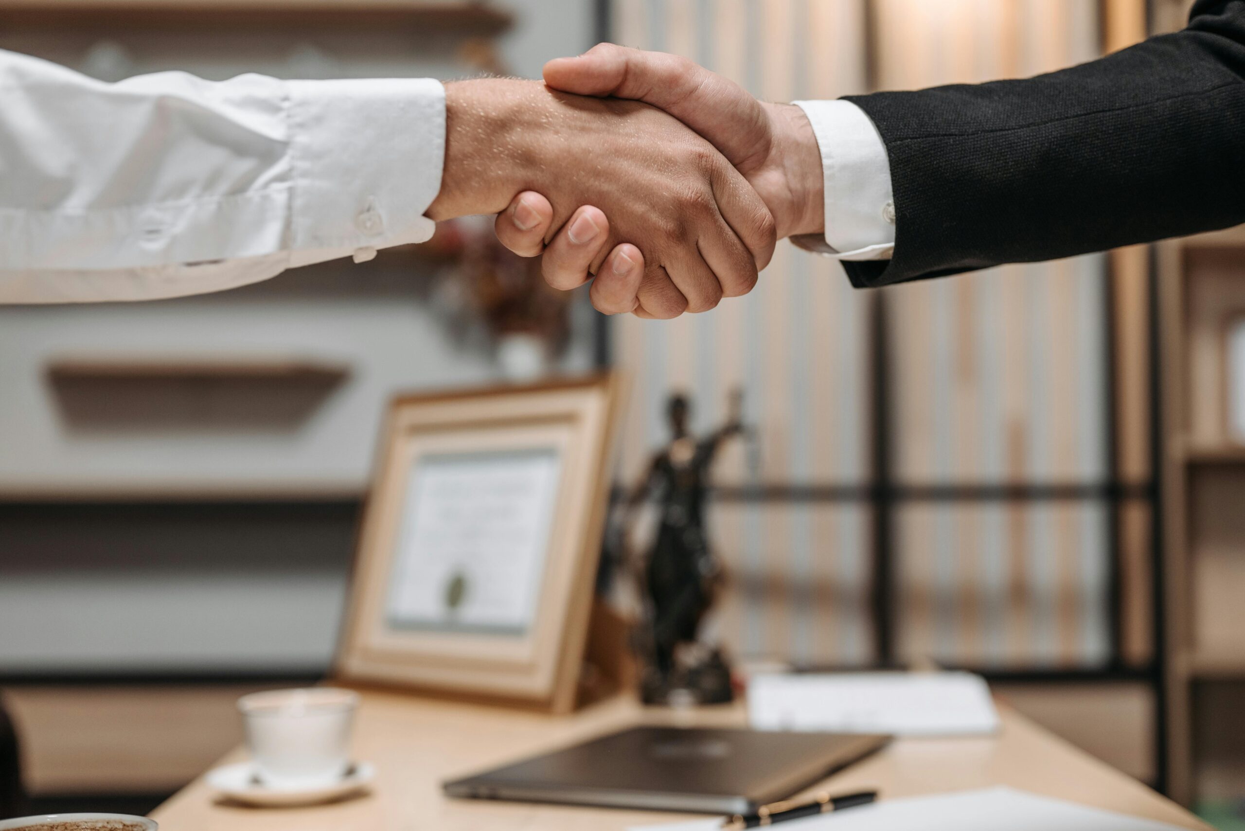 Two people in business attire shake hands across a desk in an office setting, possibly finalizing how to divide assets during divorce. A framed certificate, a small statue, a laptop, and a coffee cup are visible on the desk in the background.