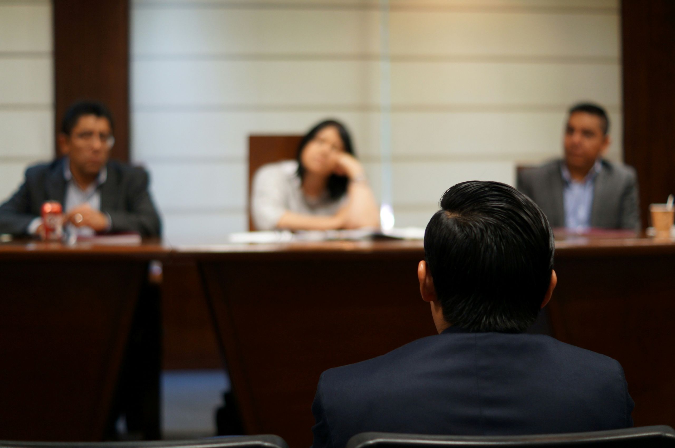 A person in a suit sits facing three people across a conference table, appearing to participate in a formal meeting or interview focused on evidence collection. The three people across the table are slightly out of focus.