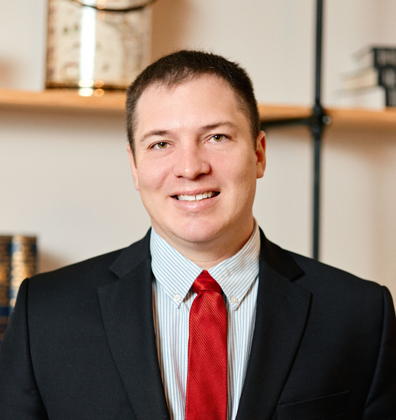 A man in a dark suit, light blue shirt, and red tie is smiling at the camera. He is standing in front of a bookshelf with books and decorative items.