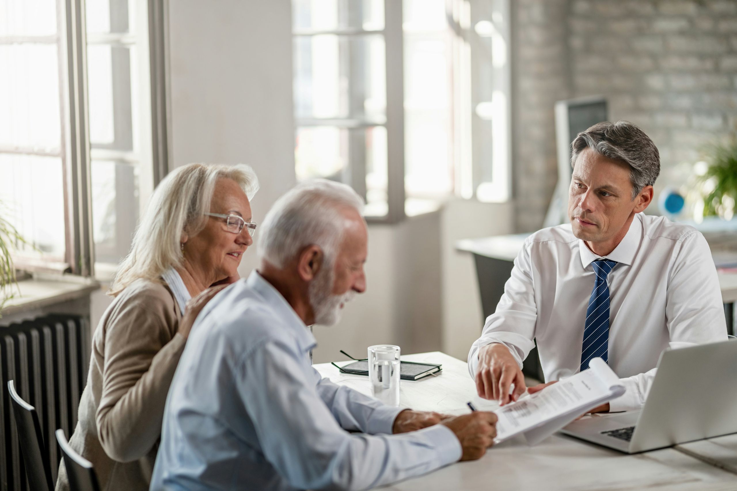 An older couple sits at a desk reviewing documents about wills vs. trusts with a professional man in a suit, who is pointing at the paperwork. A laptop and glass of water are on the table in a bright office setting.