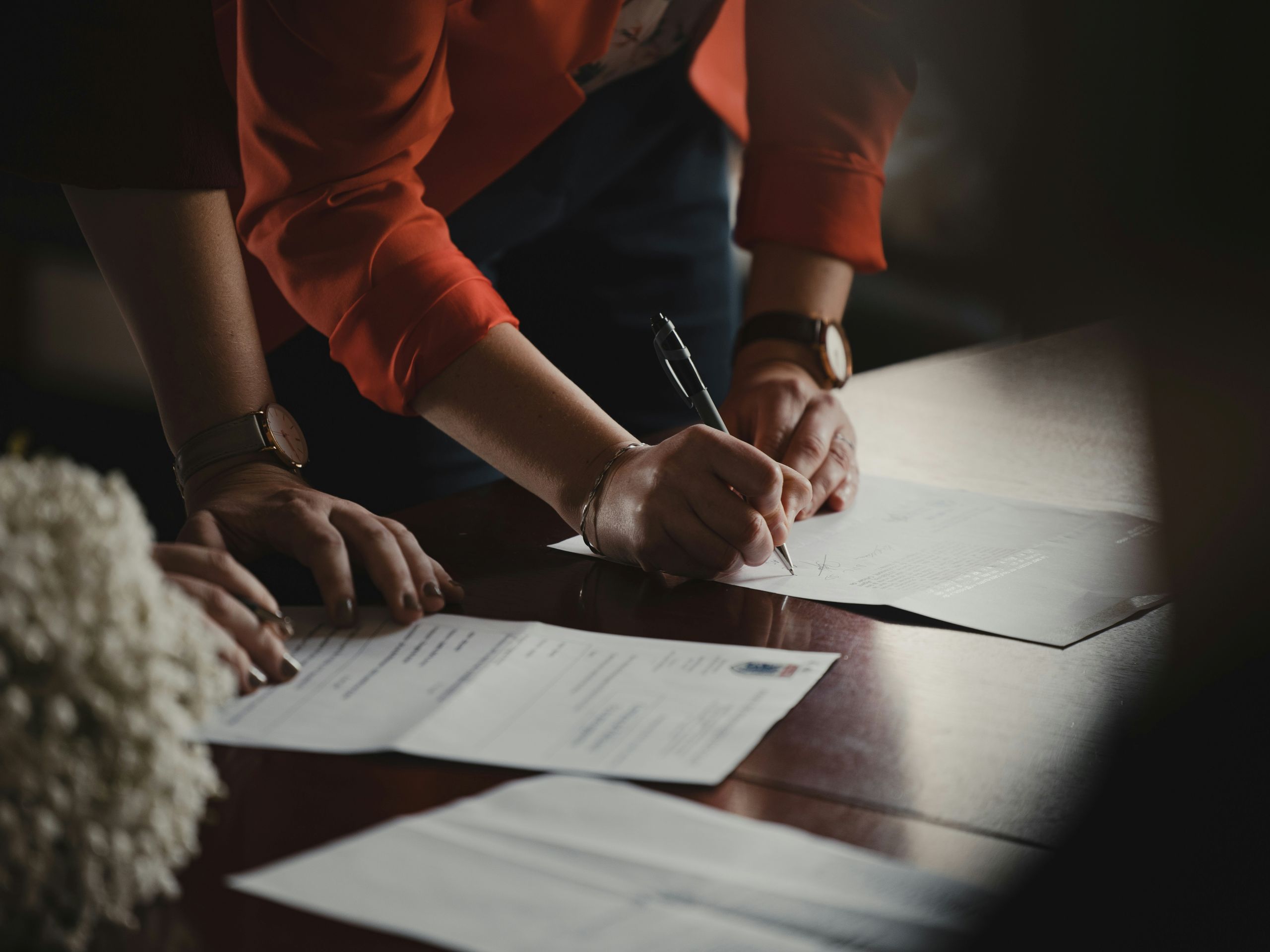 A person wearing a red jacket is signing a document on a wooden table, building trust with your lawyer, while another person stands nearby. Several papers are spread out on the table, and a bouquet of white flowers is partially visible.