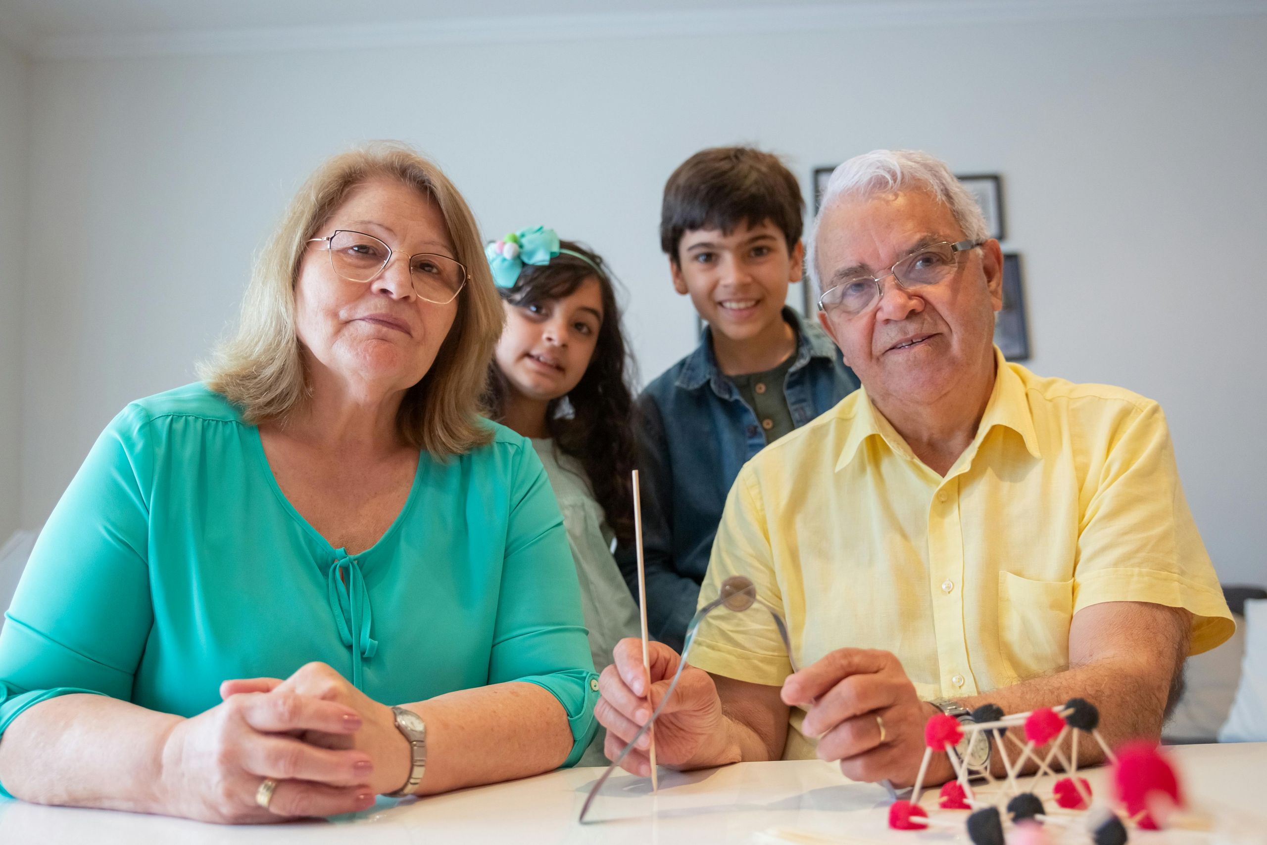 Two smiling grandparents sit at a table with two children standing behind them. As they work on a science project together, their bond highlights the importance of estate planning for securing their family's future.
