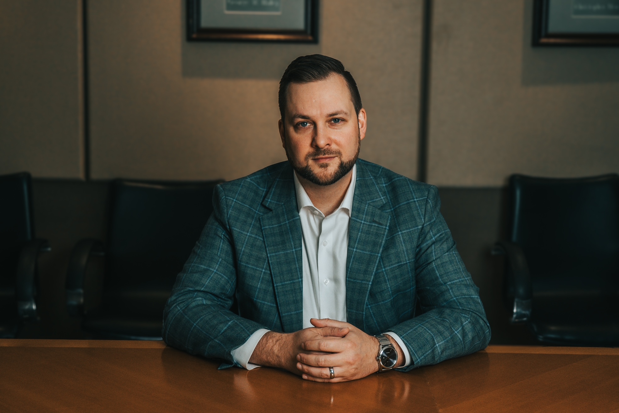 A man with short dark hair and a trimmed beard, wearing a blue plaid suit jacket and white shirt, sits at a wooden conference table with his hands folded, looking directly at the camera.