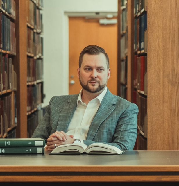 A man in a suit sits at a library table with an open book in front of him, flanked by bookshelves filled with books. He looks thoughtfully to the side.