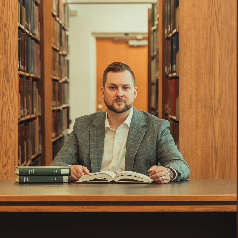A man in a suit sits at a library table with an open book in front of him and two stacked books nearby, surrounded by tall shelves filled with books.