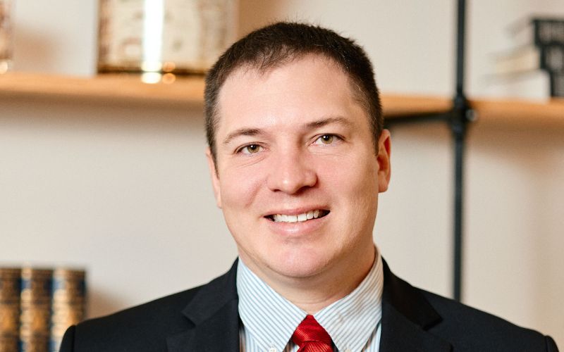 A man in a black suit, light blue shirt, and red tie smiles at the camera. He has short brown hair and is standing indoors with blurred bookshelves in the background.