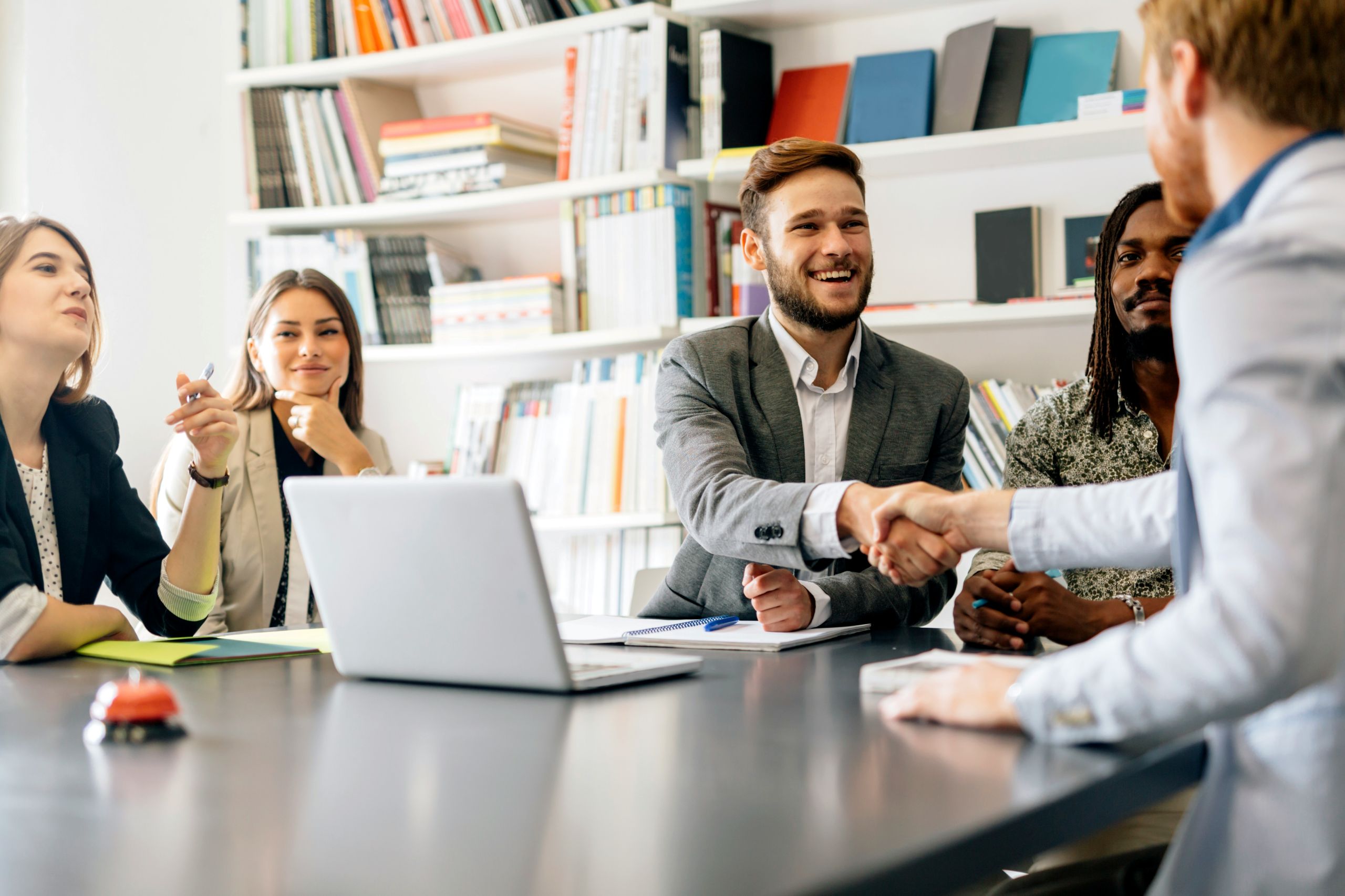 Five people sit around a conference table in a modern office, with two men—one a business lawyer—shaking hands at the center. Others look on, smiling. A laptop, papers, and books are visible in the background.