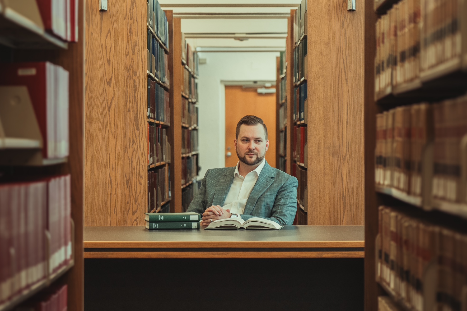 A man in a suit sits at a table in a library, surrounded by bookshelves, with an open book and two stacked books in front of him. He looks directly at the camera.