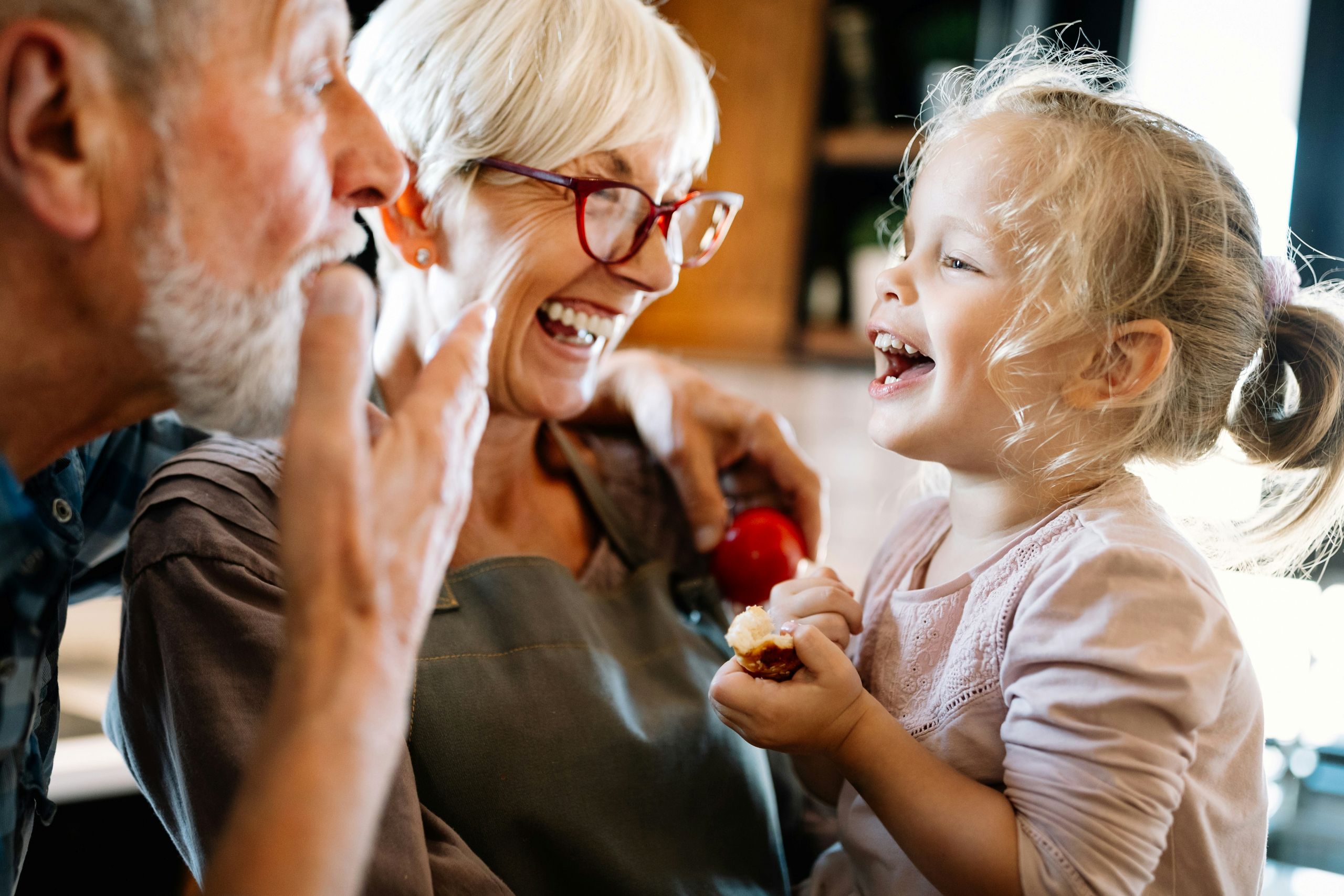 A smiling elderly couple and a young girl laugh together in a bright kitchen. The woman wears glasses and an apron, while the girl holds a piece of food, cherishing the joyful moment and the importance of grandparents’ rights.