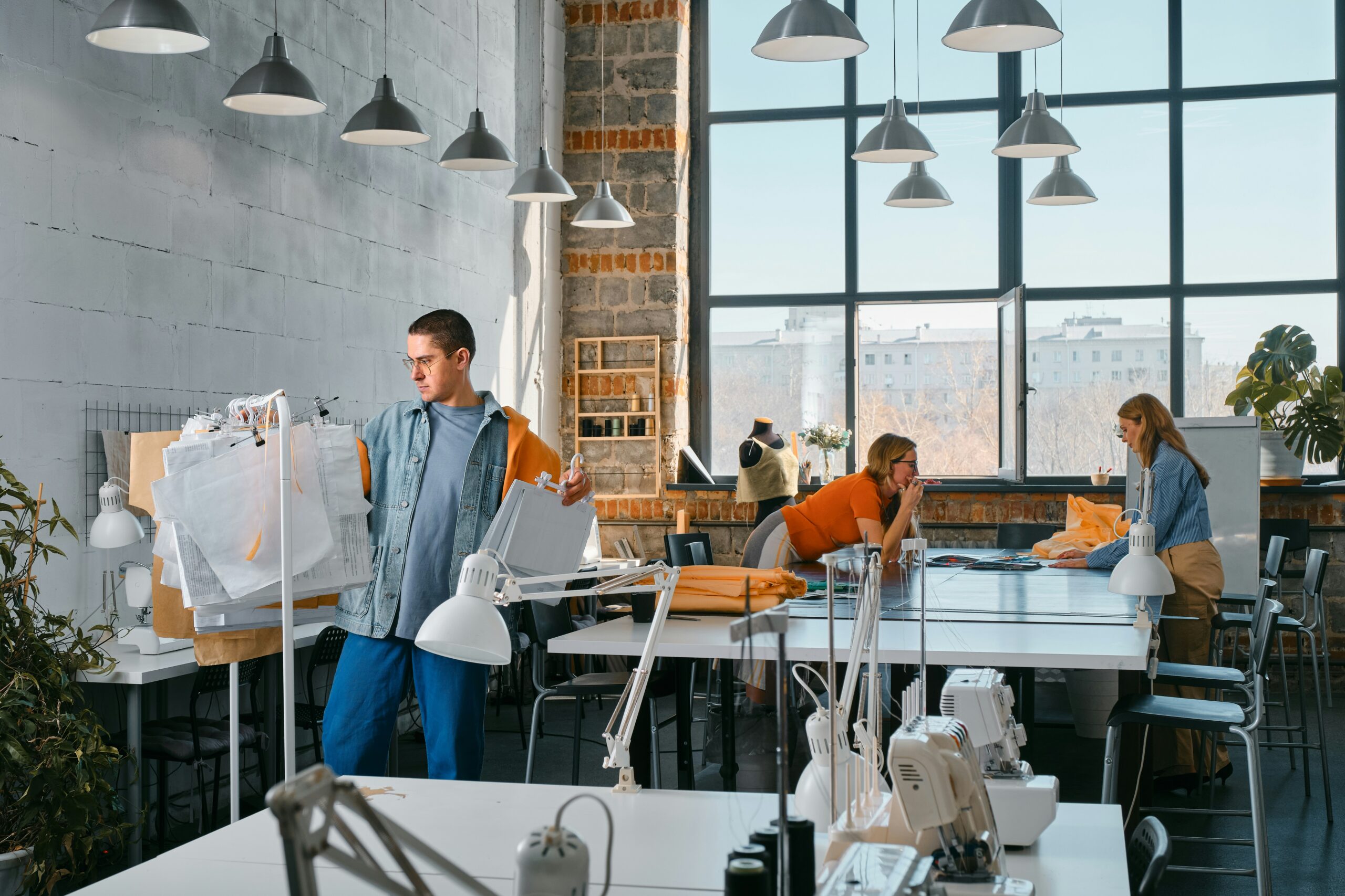 A man holds fabric patterns in a bright sewing studio, while two women work at a table—illustrating teamwork and business expansion. The space features large windows, industrial walls, sewing machines, and hanging lights.