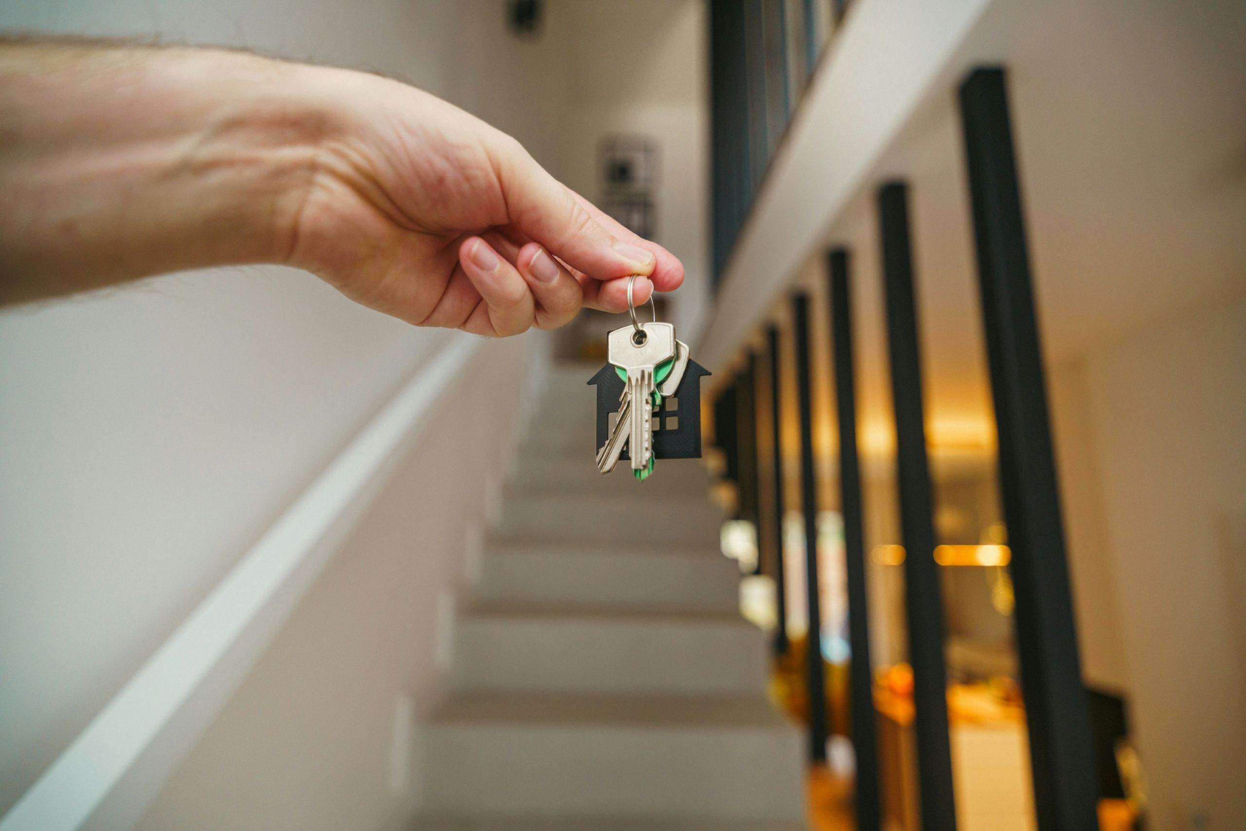 A hand holds out a set of house keys in front of a staircase inside a modern, brightly lit home, emphasizing the importance of a Legal Checklist for Landlords and Property Owners.