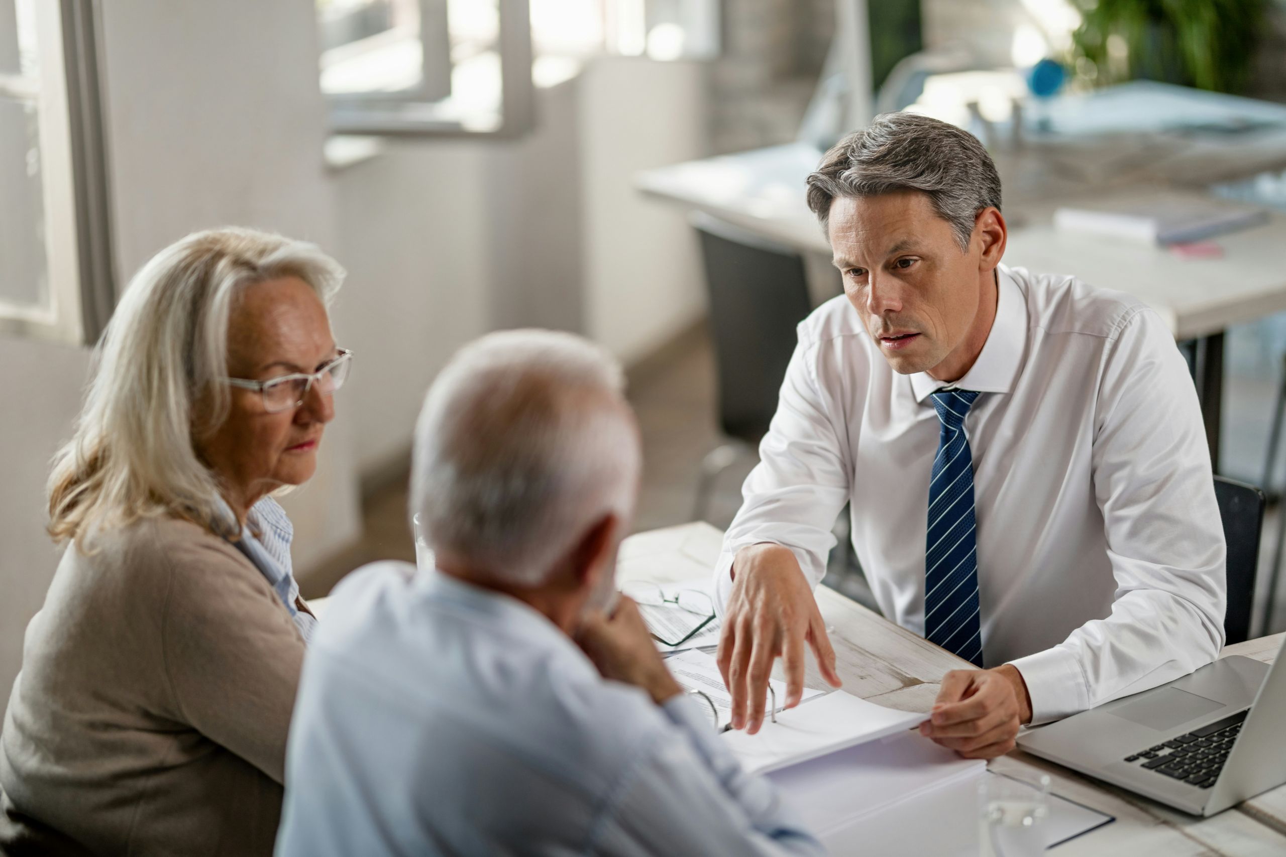 A businessman in a suit discusses strategic estate planning documents with an older man and woman at a desk in a bright office. The businessman appears focused, while the couple listens attentively. A laptop and papers are on the table.