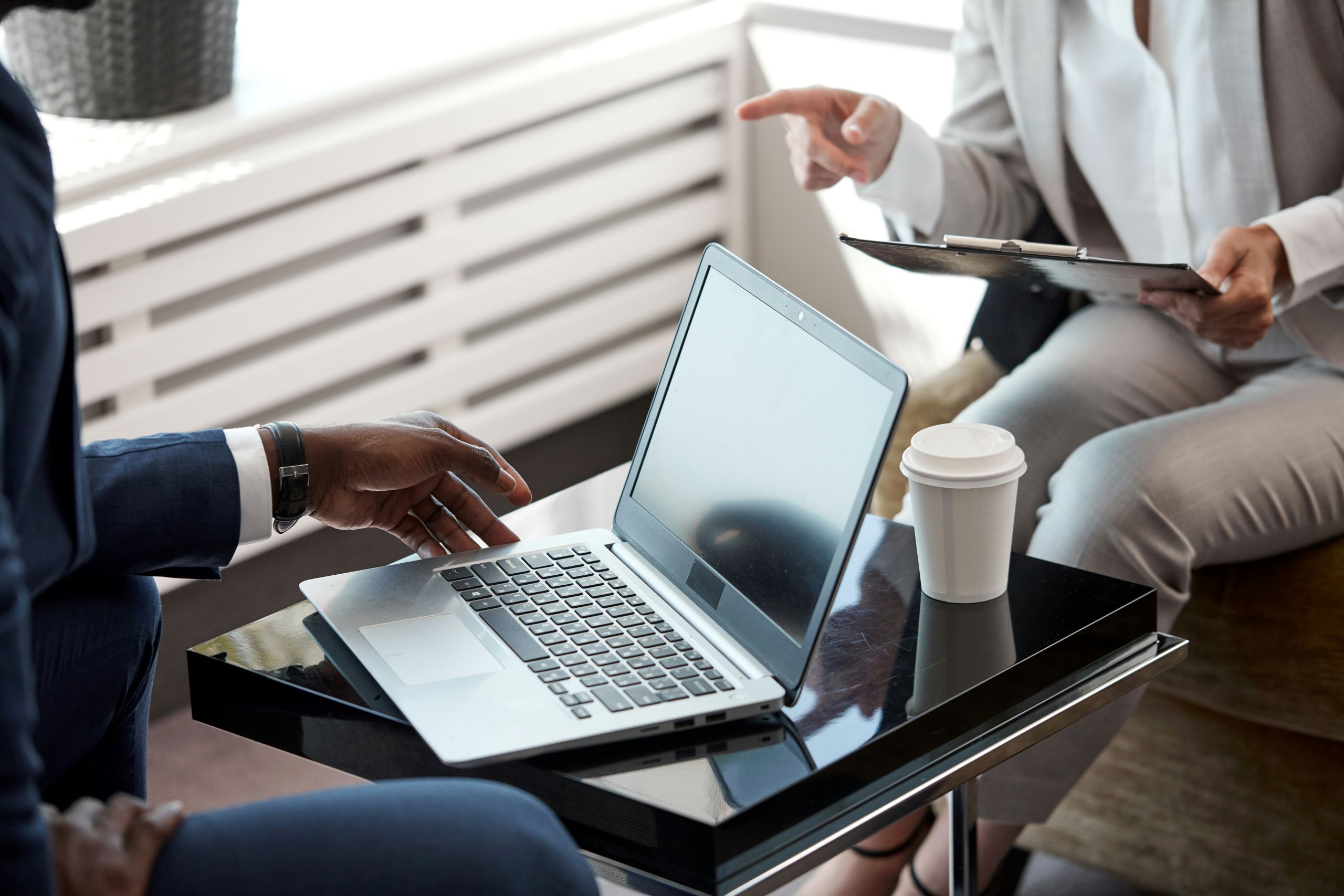 Two people in business attire sit at a table with a laptop, coffee cup, and clipboard. One gestures while the other reaches toward the laptop, suggesting a discussion or meeting with a business law attorney.