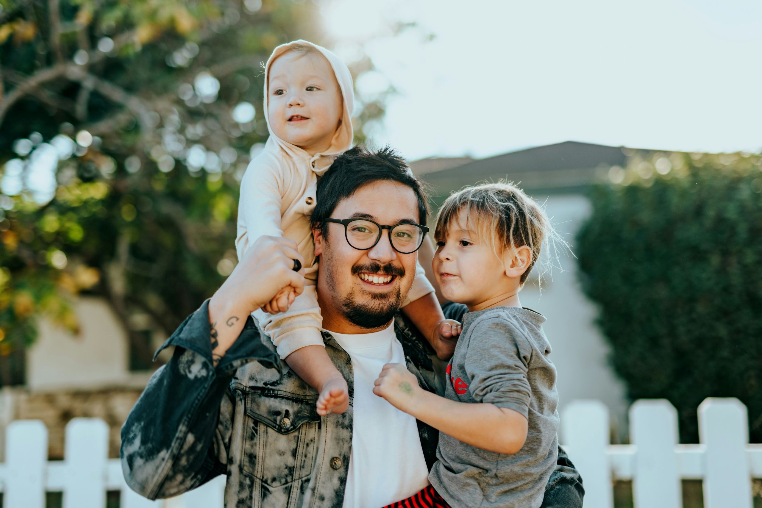 A smiling man with glasses, enjoying custody of his children, holds a baby on his shoulders and hugs a young child in front of a white picket fence, with trees and sunlight in the background.