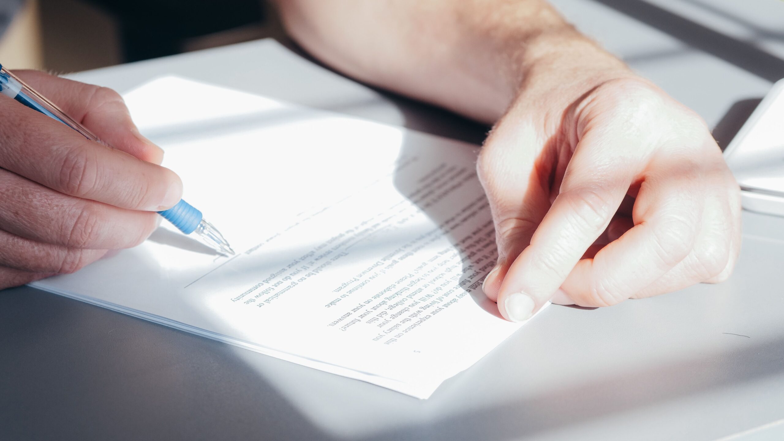 A person holding a blue pen writes on a stack of white papers on a desk, with sunlight casting shadows across their hands and the estate planning documents.